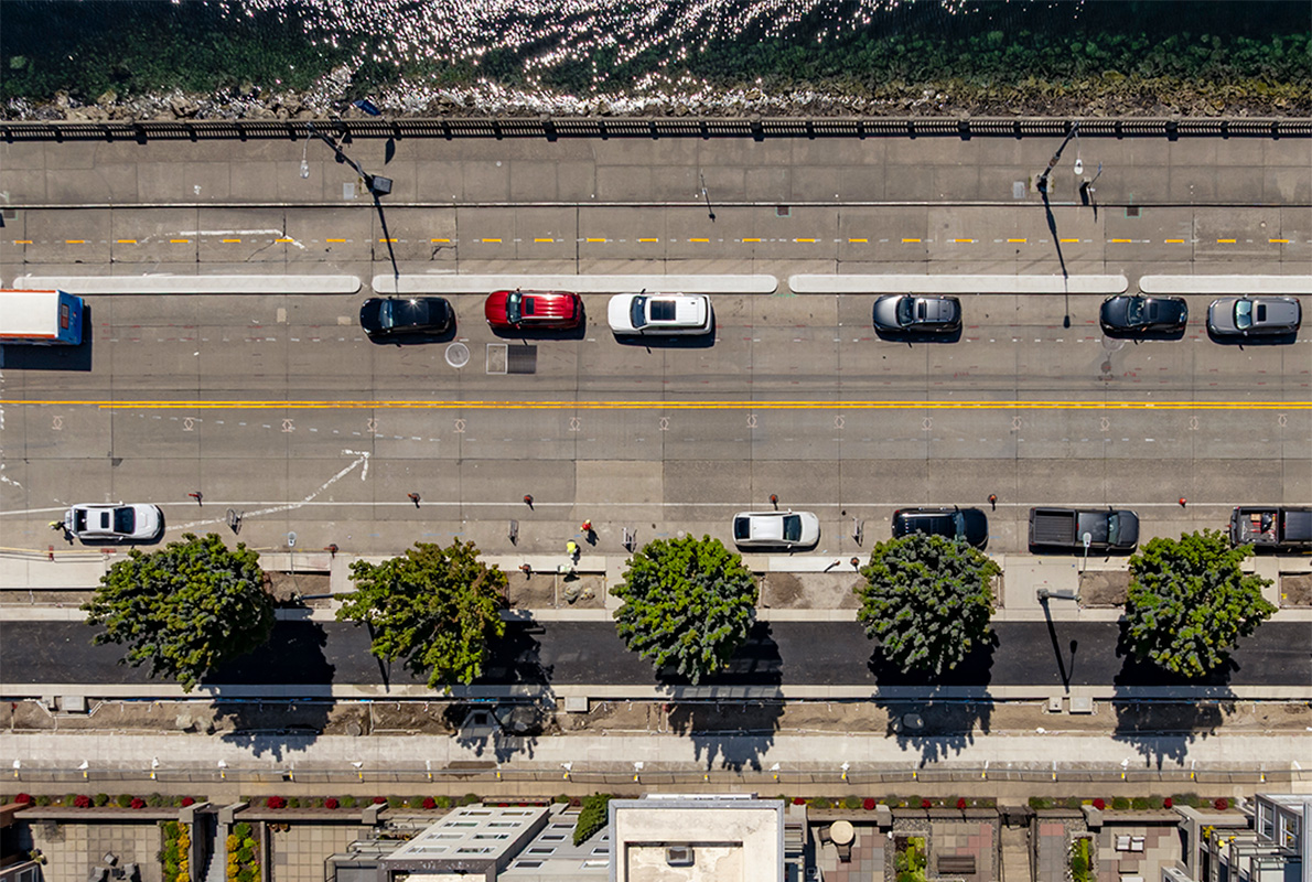 Aerial view of a waterfront city street with multiple lanes of traffic, parked cars, a bike lane, and a pedestrian sidewalk.