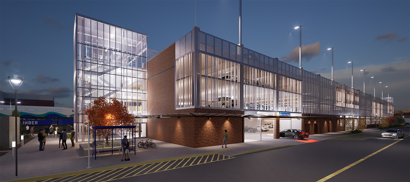 A large stairwell tower lined with windows connects to a stone parking garage with metal details and slatted windows that allow one to see inside the garage. The entire structure is brightly lit.