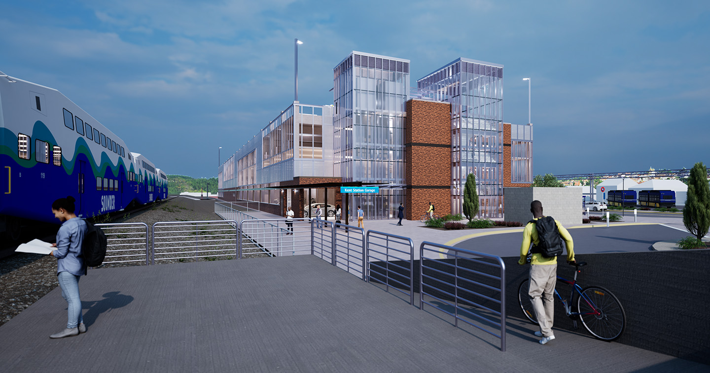 The image perspective is that of someone standing on the train platform. Two large glass stairwell towers are connected to a stone and metal parking garage. A vehicle pull-through area is in the foreground and busses can be seen parked in the background.