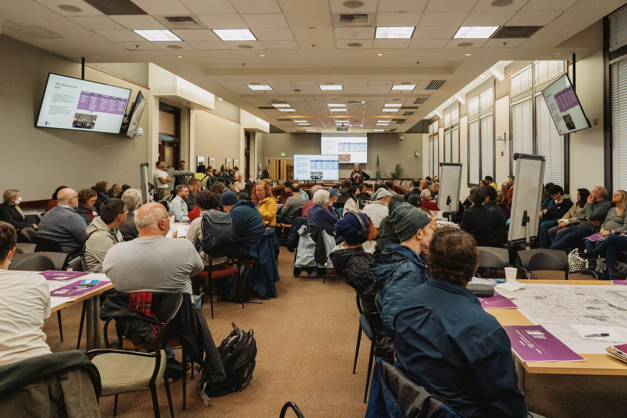 Members of the public are sitting at tables and listening to a presenter provide an overview of the SDH Workshop 1 activity.